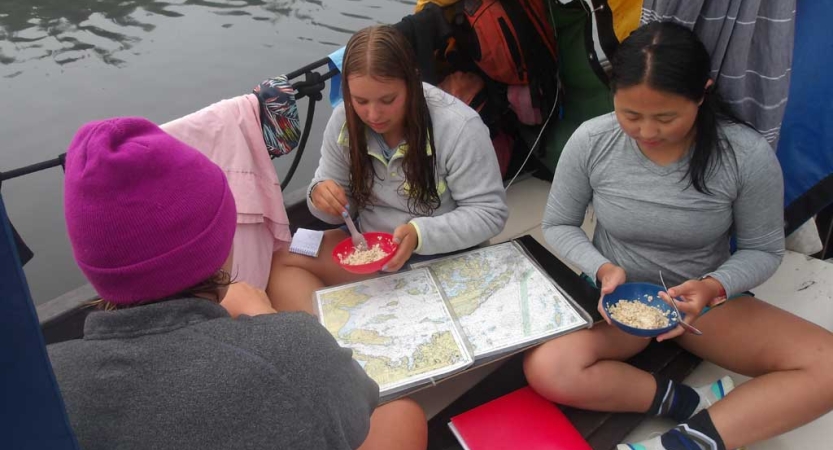 Three people sitting on a boat eat while examining a map. 